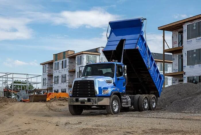 Blue Freightliner 108SD Plus dump truck in action at a bustling construction site. The truck's lifted bed is actively depositing materials, showcasing its operational capacity. In the background, multi-story residential buildings in various stages of construction highlight the site's progress. The scene captures the essential role of reliable vehicles like the Freightliner in supporting large-scale construction projects. A testament to the truck's versatility and ruggedness in demanding environments.