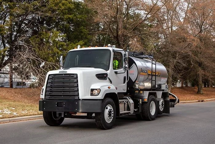 White Freightliner 108SD Plus truck designed for government and municipality applications. The vehicle is equipped with a Rosco Maximizer-3 tank, signifying its use in road maintenance or similar municipal operations. The truck is positioned on an asphalt road, flanked by mature trees in a serene, urban setting. The clear branding and specialized equipment on the truck underscore its dedication to public service tasks. This image showcases the adaptability of the Freightliner 108SD Plus to cater to specific sector needs, ensuring smooth operations in government and municipality sectors.