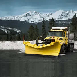 A robust Freightliner 114SD Natural Gas truck, equipped with a large snowplow, stands ready against a breathtaking backdrop of snow-covered mountains and forests. The bright yellow hue of the truck contrasts starkly with the serene winter landscape, highlighting its importance in maintaining safe road conditions in challenging terrains.