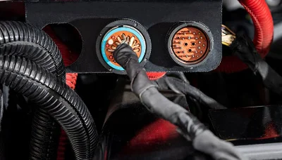 Detailed view of the Freightliner QuickFit Electrical System. The image showcases a metallic enclosure with multiple plug-in connectors, emphasizing its modular design. Surrounding wires, neatly organized, emphasize the system's easy integration and high-quality engineering. The red connector in focus suggests a primary or power connection, illustrating the user-friendly nature of the system. Ideal for highlighting advanced tech solutions in Freightliner vehicles