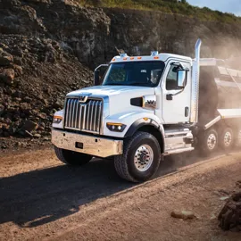A robust Western Star 47X stands prominently against a rugged backdrop. The truck, with its iconic chrome grille and sturdy design, is evidently built for tough terrains, as it navigates through a construction or mining site with ease. Dust rises from the ground as the truck hauls material, emphasizing its heavy-duty capabilities. The white paint and distinctive branding reflect the sunlight, drawing attention to the vehicle's meticulous craftsmanship and reliability.