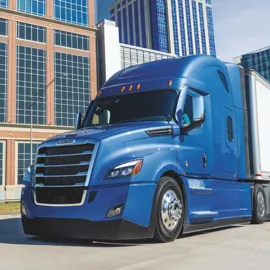 Blue Freightliner Cascadia truck driving through an urban area with modern office buildings in the background, showcasing efficient city transportation.