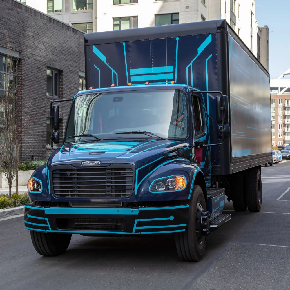 Freightliner eM2 electric truck driving through urban streets with modern buildings and blue graphic design on black trailer