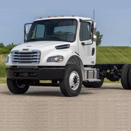 White Freightliner M2 106 chassis parked on a paved road with a lush green backdrop, highlighting its robust design and versatility.
