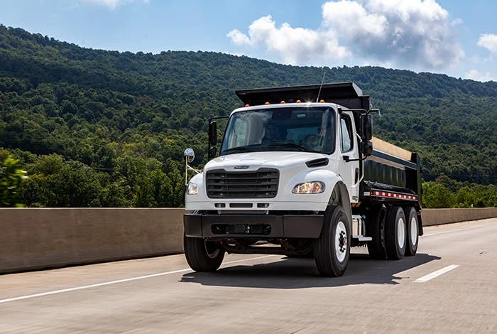 Freightliner M2 106 dump truck driving on a highway with scenic green mountains in the background