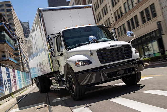 Freightliner M2 106 delivery truck driving through a city street with tall buildings