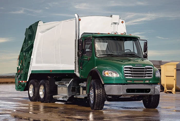 Freightliner M2 106 Plus green garbage truck parked on a wet surface with clear sky