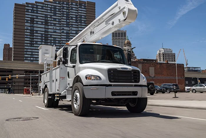 Freightliner M2 106 Plus utility truck with extended white boom driving through a city with tall buildings