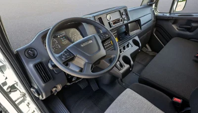 Intimate view of the Freightliner 108SD Plus truck's interior, highlighting its ergonomic design and spacious cabin. The driver's area is well-equipped with a modern steering wheel featuring control buttons, and a clearly organized dashboard. The black and gray color scheme of the seats and dashboard ensures a professional and sleek appearance, while the fabric seats promise comfort for long drives. This interior showcases the blend of functionality and comfort that professionals can expect from the 108SD Plus.