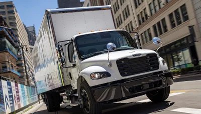 Freightliner M2 106 Plus truck driving through a city street with a white cargo box