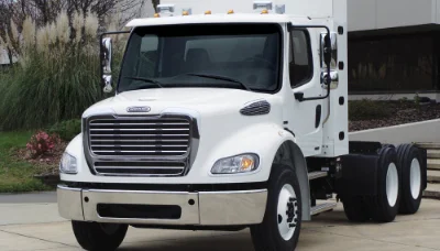 Frontal view of a white Freightliner M2 112 Natural Gas truck cab chassis parked in a landscaped area near a building