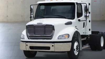 Frontal view of a white Freightliner M2 112 Natural Gas truck cab chassis parked in a landscaped area near a building