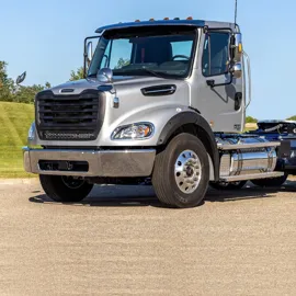 Silver Freightliner M2 112 truck standing on a paved road, highlighting its sturdy build, chrome accents, and detailed craftsmanship against a clear sky backdrop.