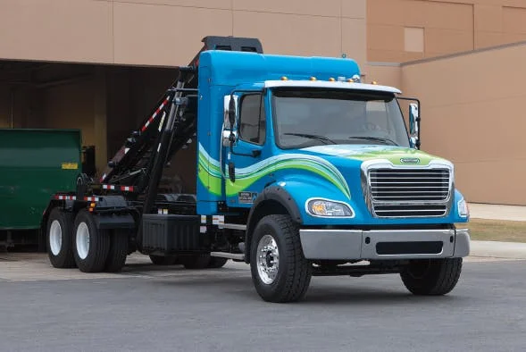 Freightliner M2 112 Plus Series in blue with green stripes, equipped with rear loader near a building