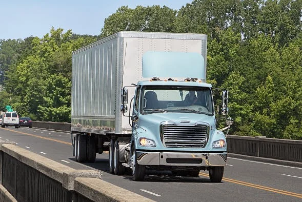 Freightliner M2 112 Plus Series in light blue transporting goods on a highway bridge surrounded by trees